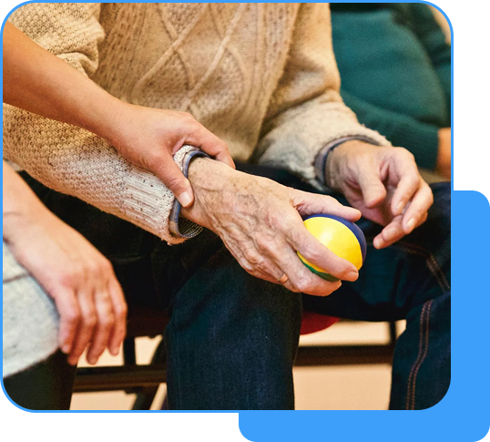 A caregiver holding the hand of an elderly person with a yellow and blue ball, sitting together in a supportive setting.