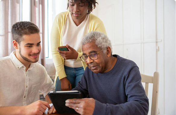 An elderly man with gray hair using a tablet with a young man and a woman in a yellow cardigan assisting, in a bright room.