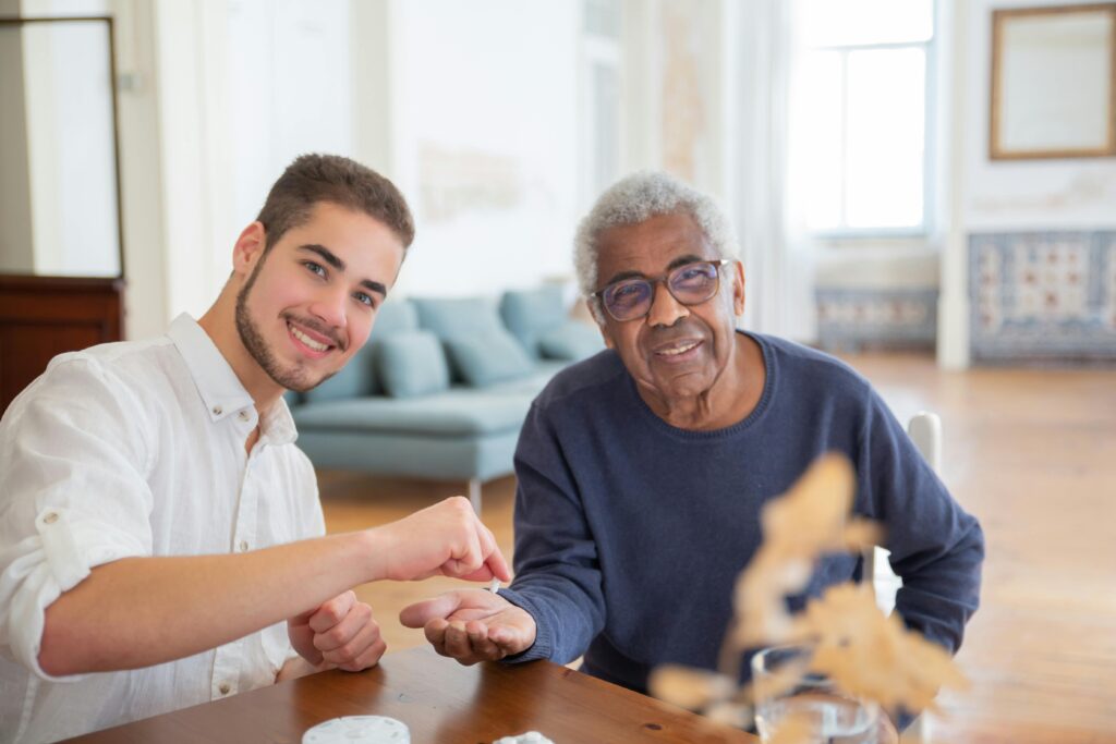 A young man in a white shirt handing something to an elderly man with gray hair in a blue sweater, sitting at a wooden table in a bright room.