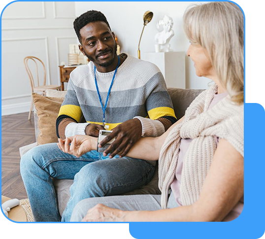 A caregiver in a colorful sweater with a lanyard checking the pulse of an elderly woman with white hair on a couch in a cozy room.