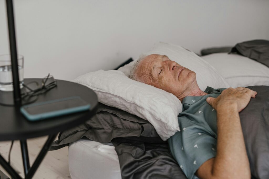 An elderly man with white hair sleeping peacefully in bed, with a bedside table holding a glass of water, glasses, and a phone.