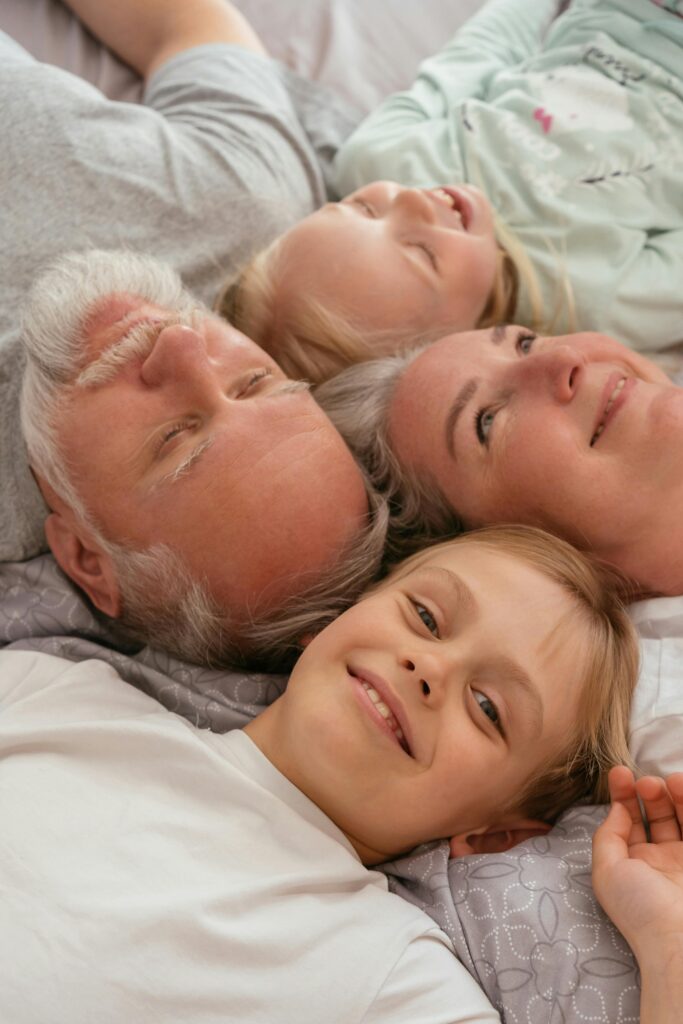 A family with an elderly man, a woman, and two children smiling and resting together on a bed in a cozy bedroom.