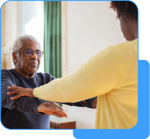 A caregiver in a yellow sweater assisting an elderly man with gray hair in a blue sweater, guiding his arms in a bright room.