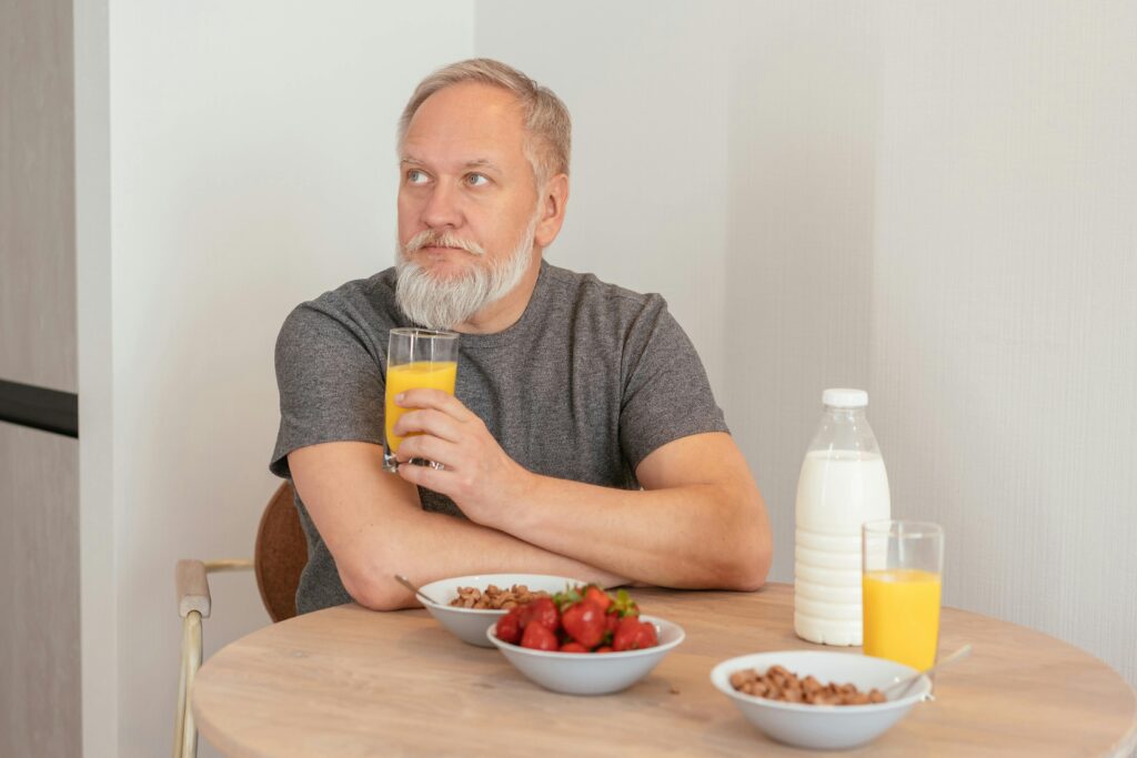 An elderly man with a white beard holding a glass of orange juice at a table with bowls of strawberries, cereal, milk, and another glass of orange juice.