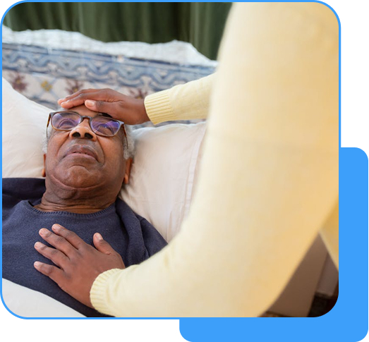 A caregiver in a yellow sweater gently placing a hand on the forehead of an elderly man with gray hair lying in bed.