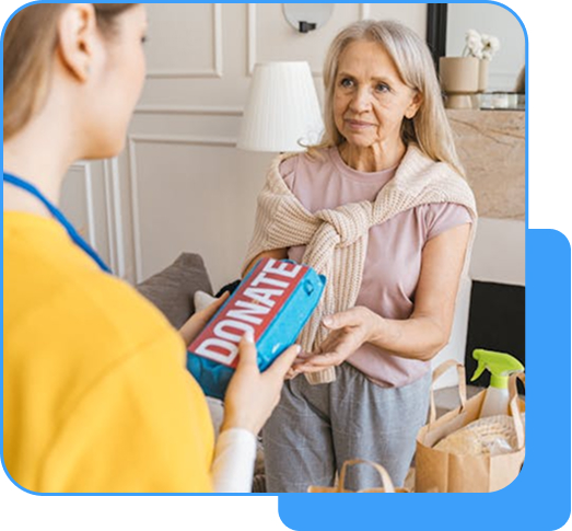 A caregiver in a yellow uniform handing a "DONATE" box to an elderly woman with white hair in a pink top, with bags and cleaning supplies nearby.